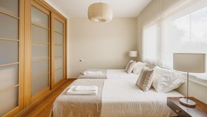 A bedroom with two beds, a bedside table with a lamp, a large window with sheer curtains, and a wooden wardrobe with frosted glass panels. - Sunny Central Luxury, 2 bedroom/2WC, garage, don't need car, 8 min walk beach (Cascais)