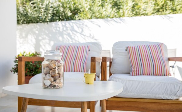 A round white table with a glass jar of seashells and a yellow cup. Two white chairs with colorful striped pillows are in the background.