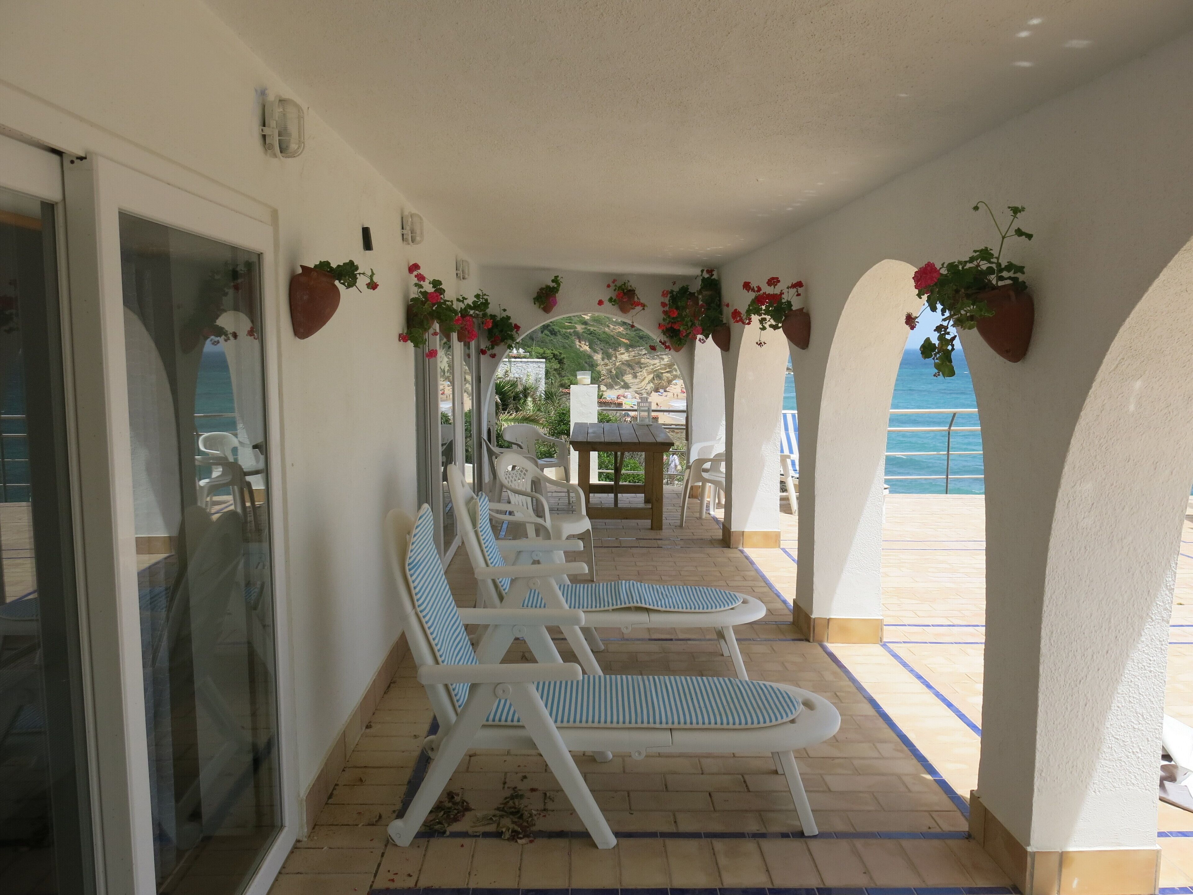 A covered outdoor seating area with white lounge chairs and tables, potted plants, and a view of the sea.
