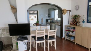 Ein Wohnzimmer mit Fernseher, Holzstühlen, einem Esstisch, einer durch einen Bogen sichtbaren Küche, einem Weinkühlschrank und einer Wanduhr.