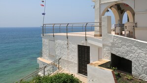 Ein Küstengebäude mit Balkon und Meerblick, mit einer Steinfassade, einer schwarzen Tür und einer kleinen Sitzgelegenheit im Freien.
