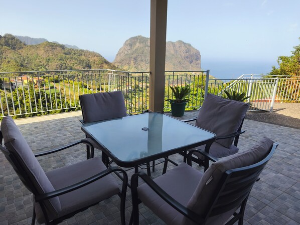 A balcony with a glass table and four cushioned chairs, overlooking a mountainous landscape.