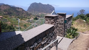 A stone staircase leading to a balcony with a metal railing, overlooking a coastal landscape with houses and a mountain in the distance.