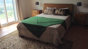 A bedroom with a bed, two nightstands, a green throw blanket, patterned pillows, a wooden headboard, and a view through a large window.