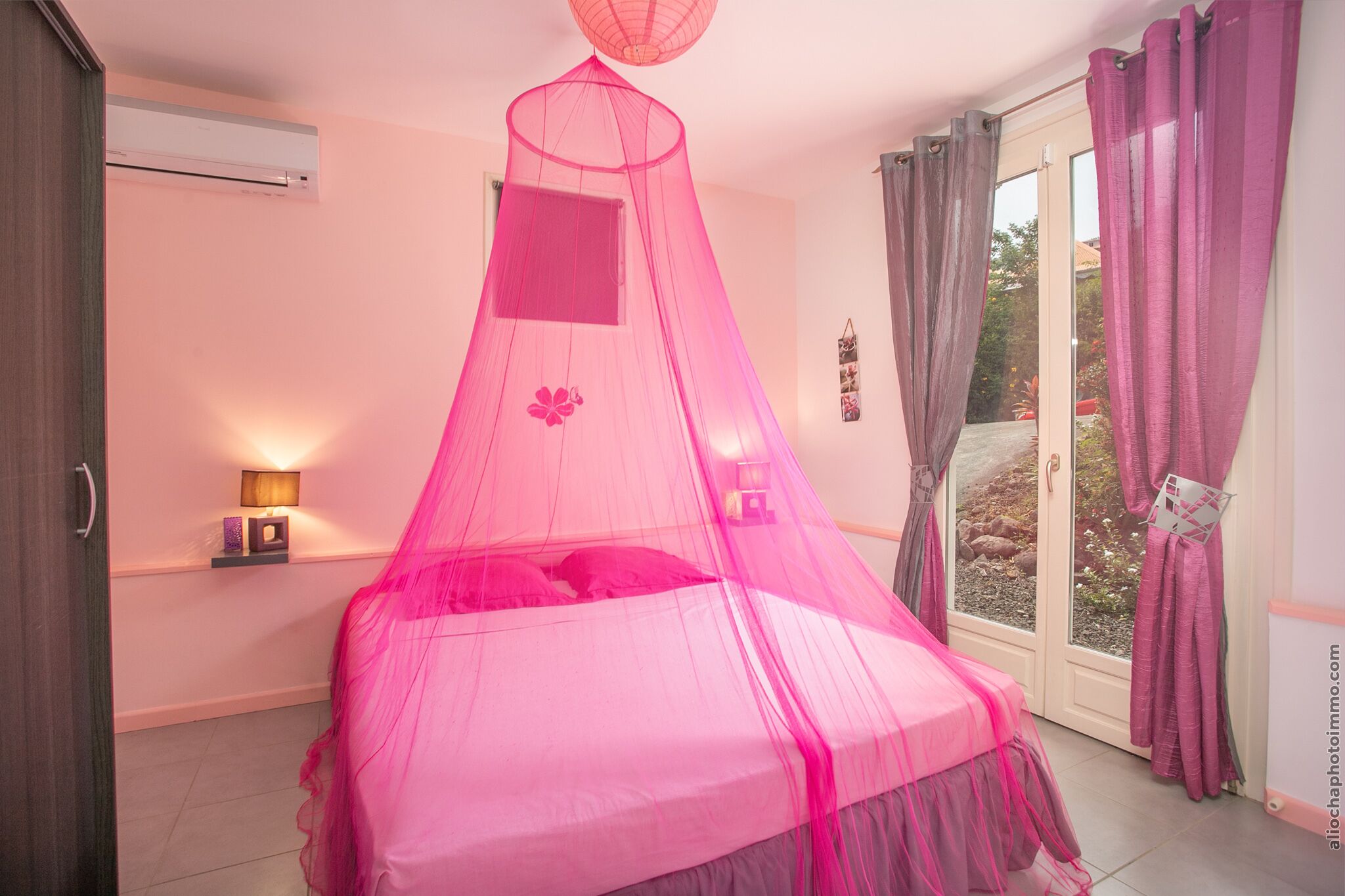 A bedroom with a bed covered by a pink canopy, a bedside table with a lamp, and a window with curtains.