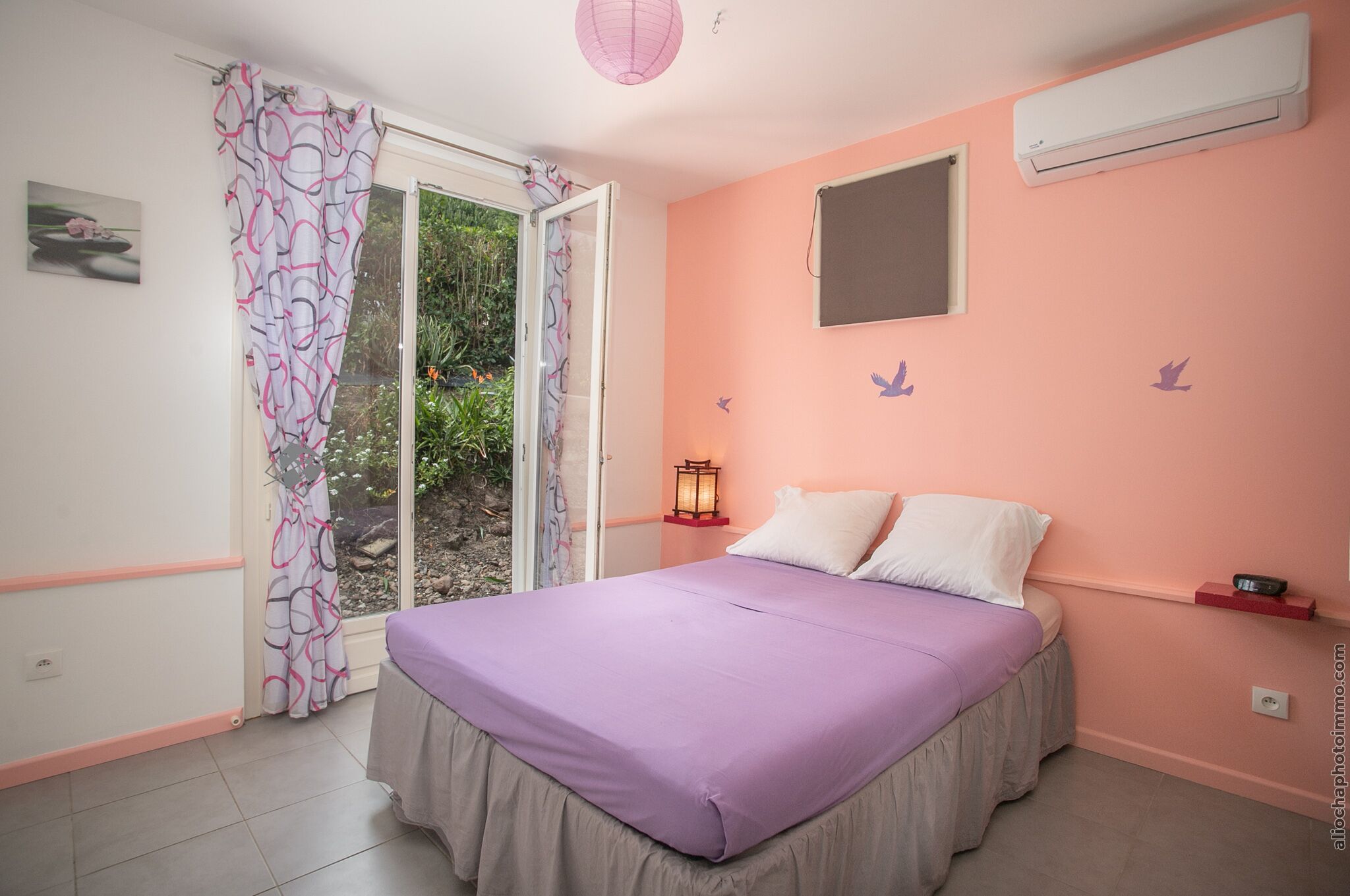 Ein Schlafzimmer mit einem Bett, einem Fenster mit Vorhängen, einer Klimaanlage und einem an der Wand befestigten Regal.