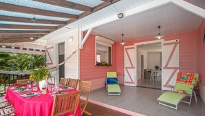 Un patio couvert abrite une table à manger pour quatre personnes, entourée de chaises en bois. On y trouve également deux fauteuils et une petite table sur laquelle est posée une plante. Les murs sont peints en rose, et une porte donne accès à une autre pièce.