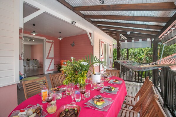 An outdoor dining area with a long table set for a meal, surrounded by wooden chairs and a covered patio.