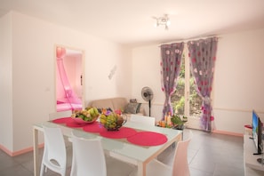 A dining area with a glass table, white chairs, a bowl of fruit, and a fan.