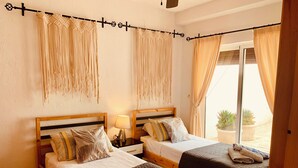 Habitación de hotel con dos camas, cabecera de madera, mesita de noche con lámpara y vista al exterior a través de una puerta corrediza de vidrio.