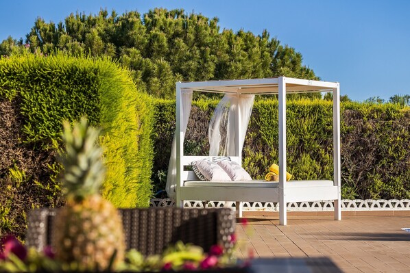 Ein weißes Himmelbett mit weißem Baldachin, platziert auf einer Holzterrasse mit Hecke und Bäumen im Hintergrund.