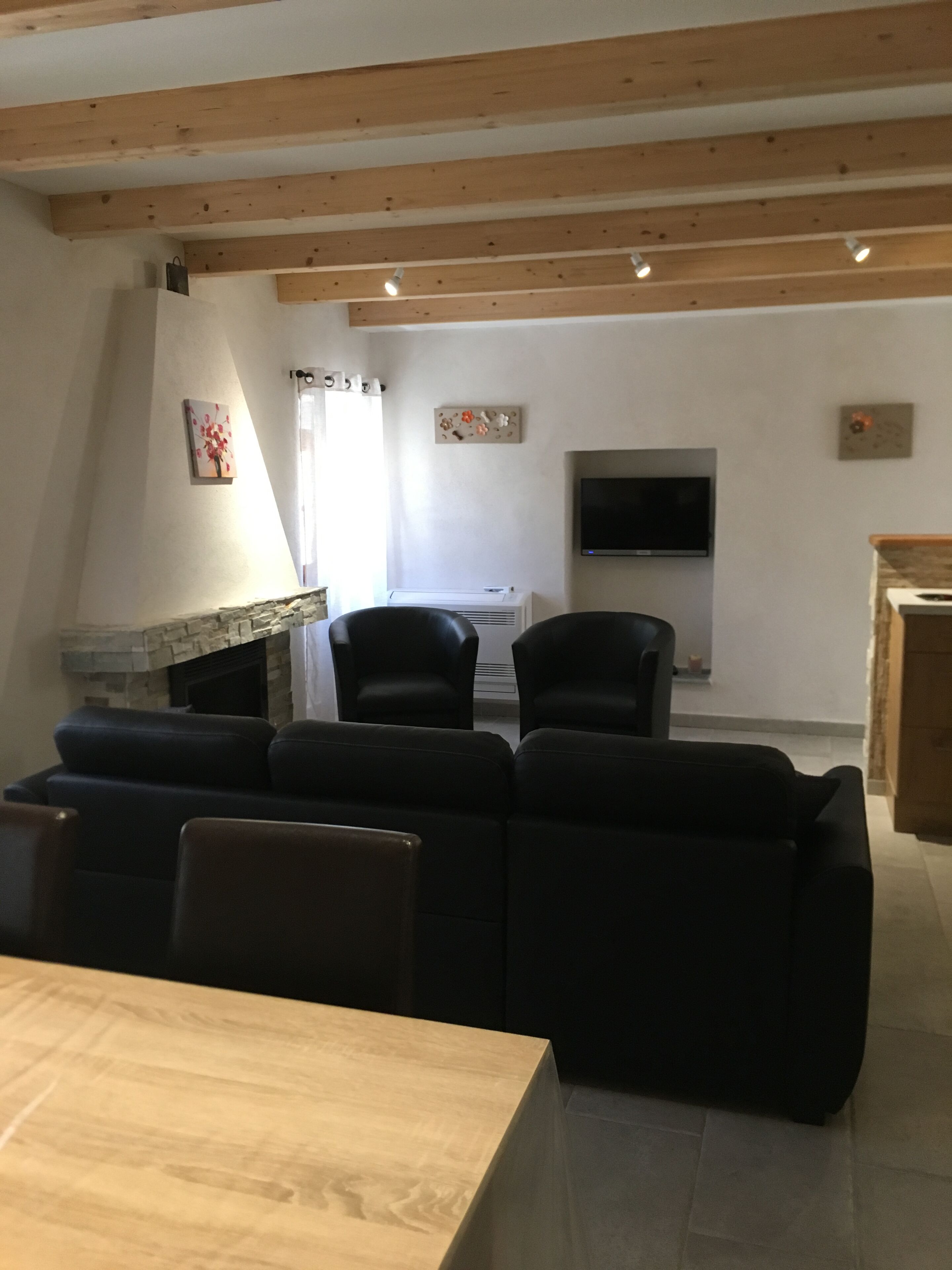 A modern living room with a stone fireplace, black leather sofas, a wooden dining table, and a flat-screen TV mounted on the wall.
