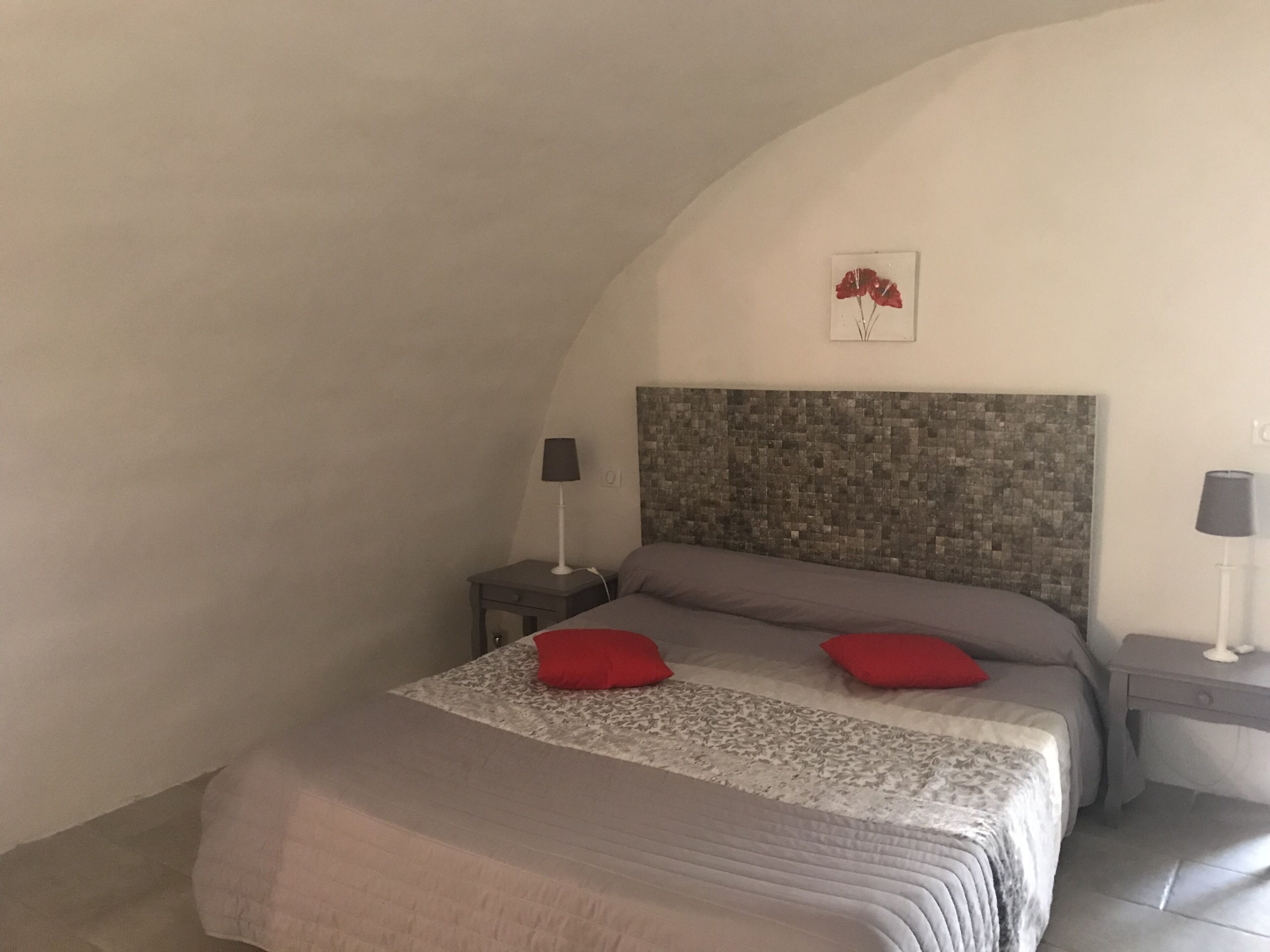 A bedroom with a sloped ceiling, a bed with a patterned bedspread, two red pillows, a headboard with a mosaic design, and two bedside tables with lamps.