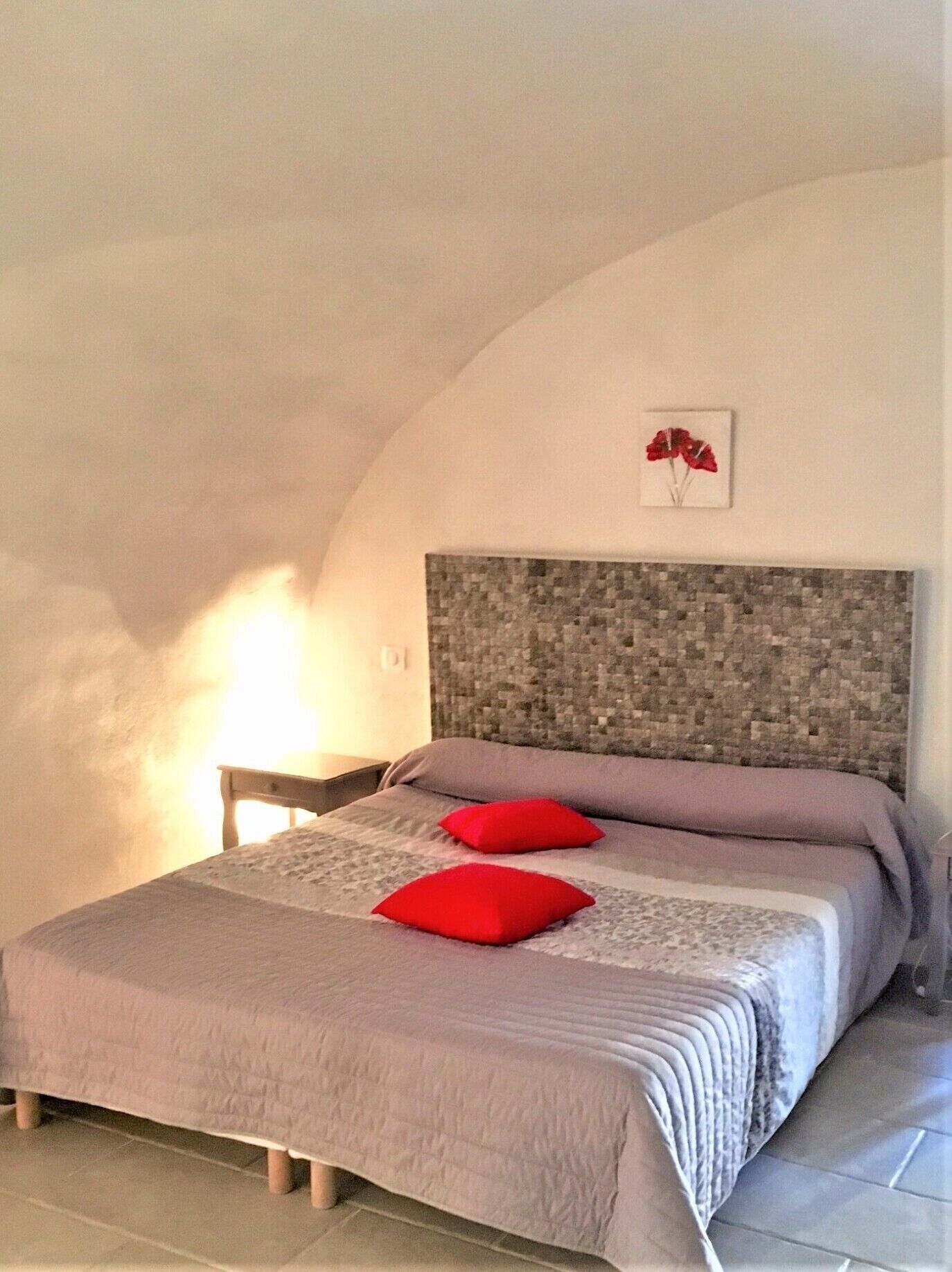 Ein Schlafzimmer mit einem steinmosaik-Kopfende, einem Bett mit grauer Steudecke und roten Kissen sowie einem kleinen Nachttisch mit Lampe.
