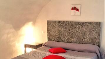 Ein Schlafzimmer mit einem steinmosaik-Kopfende, einem Bett mit grauer Steudecke und roten Kissen sowie einem kleinen Nachttisch mit Lampe.