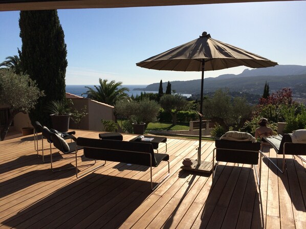 Eine Holzterrasse mit Gartenmöbeln und einem großen Sonnenschirm, mit Blick auf eine malerische Landschaft mit Bergen und einem Gewässer.