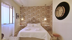 A neatly made bed with white linens, two bedside tables with lamps, a round mirror on the wall, and a window with curtains.