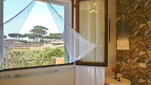 A room with a view of trees and a building through a window with a white curtain. There is a small table with a vase on it.