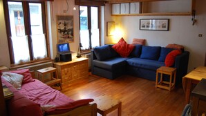 A cozy living room with a blue sofa, a red chair, and a wooden table. There is a window with blinds, a lamp, and a small stool. - Apartment in Beautiful Alpine Village (Chamonix-Mont-Blanc)
