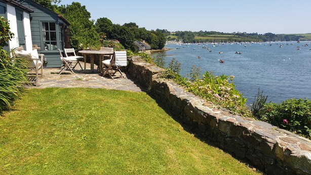 A lakeside property with a stone wall, a grassy lawn, and a wooden deck with chairs. - Beautiful cottage with sea views and private beach access (Helston)