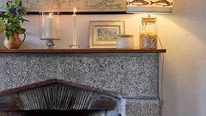 A stone fireplace with a lit fire, a map on the wall, a lamp with fish patterns, and a basket of wood. - Beautiful cottage with sea views and private beach access (Helston)