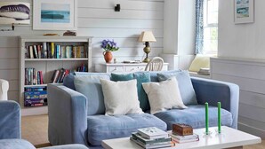A living room with a blue sofa, a white coffee table, and a bookshelf filled with books. - Beautiful cottage with sea views and private beach access (Helston)