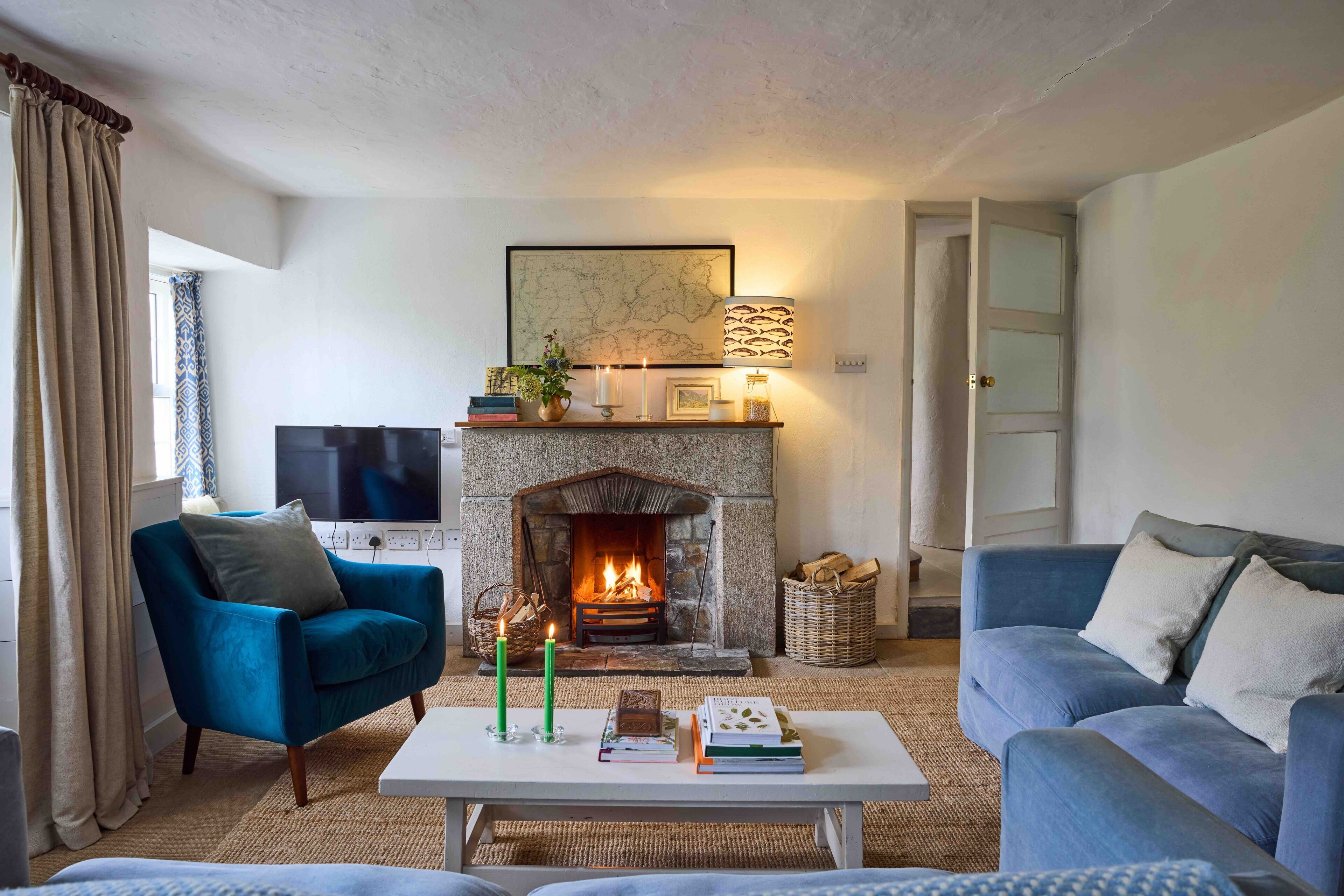 Ein Wohnzimmer mit Kamin aus Stein, einem blauen Sessel, einer Couch, einem Flachbildfernseher und einem Couchtisch mit Büchern und Kerzen.