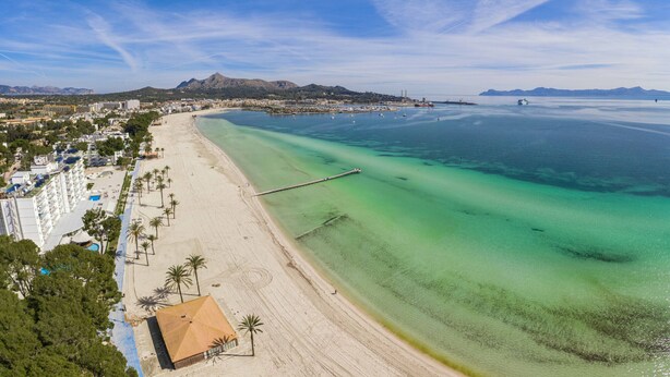 Ein Küstenstrand mit klarem türkisblauem Wasser, einem langen Sandstrand und einem kleinen Gebäude mit rotem Dach.