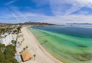 A coastal beach with clear turquoise waters, a long sandy shore, and a small building with a red roof. - Villa Diana (Puerto de Alcudia)