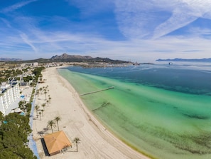 A coastal beach with clear waters, a long pier, and a backdrop of mountains.