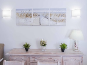 A room with a white wall, a wooden dresser, two potted plants, a vase with flowers, and a painting above the dresser.