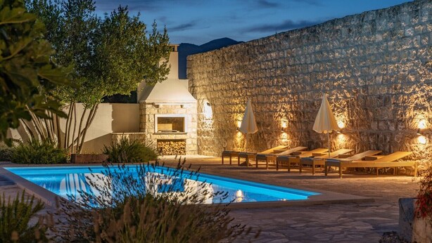 A swimming pool with lounge chairs and umbrellas, a stone wall, and a fireplace.