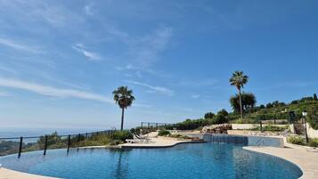 Une piscine à bord incurvé, entourée d’une terrasse carrelée et d’un mur en pierre. On aperçoit des palmiers et un paysage en arrière-plan.