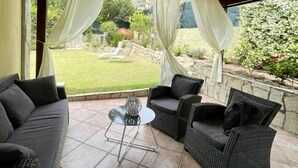 Une terrasse extérieure avec un canapé noir, deux fauteuils en osier et une petite table ronde.