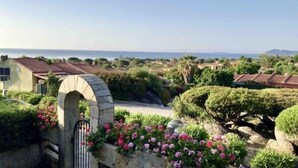 Un arc en pierre mène à un jardin où fleurissent des plantes aux couleurs roses, et d’où l’on peut admirer un paysage côtier.