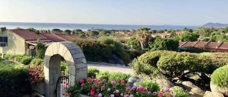 Un arco de piedra que da acceso a un jardín con flores rosadas y vistas al paisaje costero.