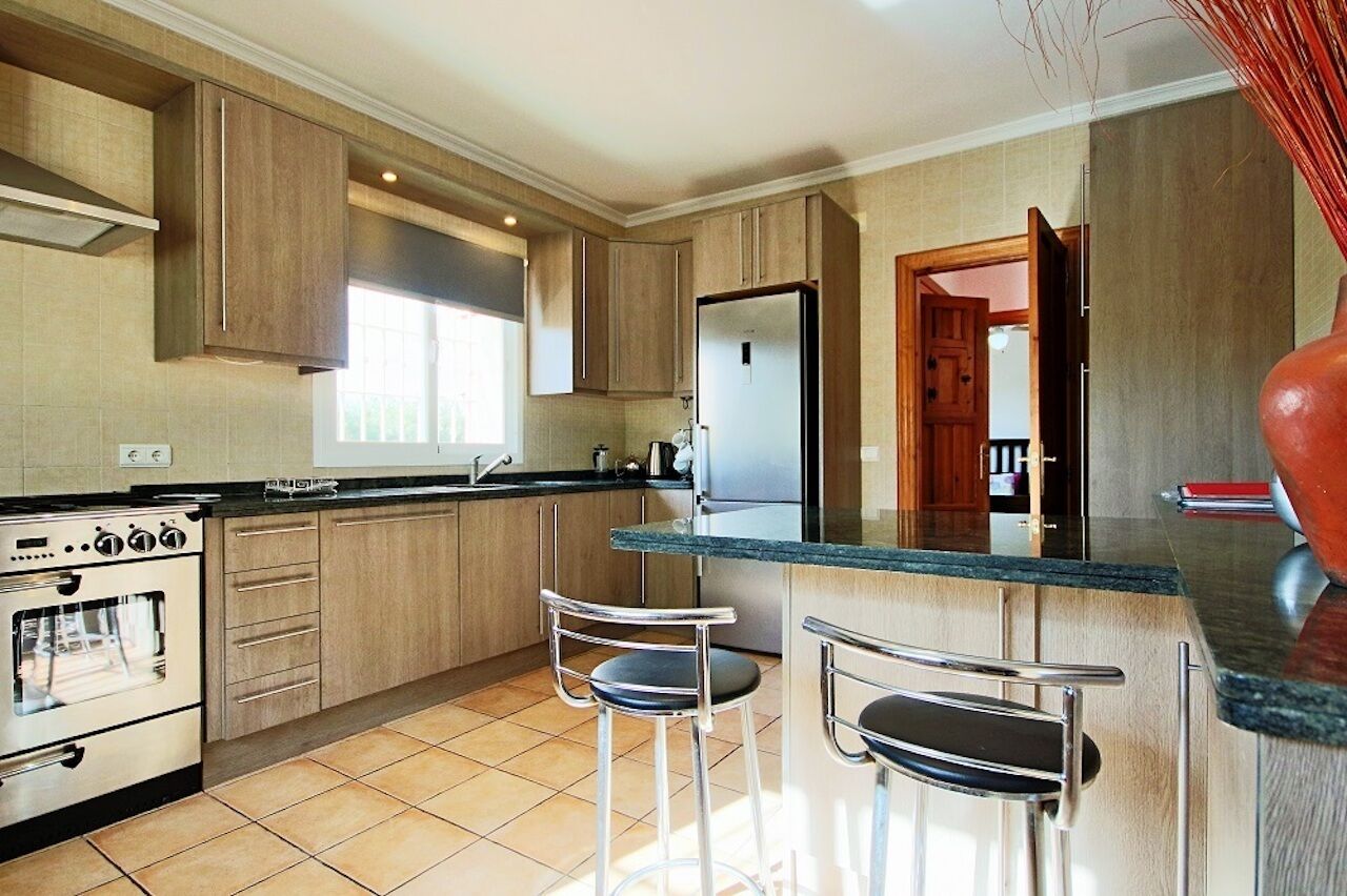 A modern kitchen with wooden cabinets, a stainless steel refrigerator, a built-in oven, and two bar stools.