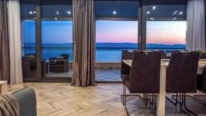 A modern dining area with a view of the sea at sunset, featuring a wooden herringbone floor, dark upholstered chairs, and a glass door leading to an outdoor seating area. - Luxury Villa POCRNJA with private heated pool, jacuzzi, sauna and pool table (Stanici)