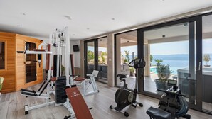 A modern gym with a variety of equipment, large windows, and a view of a pool and mountains. - Luxury Villa POCRNJA with private heated pool, jacuzzi, sauna and pool table (Stanici)