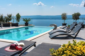 An infinity pool with lounge chairs and potted plants, overlooking a body of water and distant hills. - Luxury Villa POCRNJA with private heated pool, jacuzzi, sauna and pool table (Stanici)