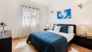 Hotellihuone, jossa on sänky, kaksi yöpöytää valaisimineen, taulu seinällä ja ikkuna, jossa on läpikuultavat verhot.
