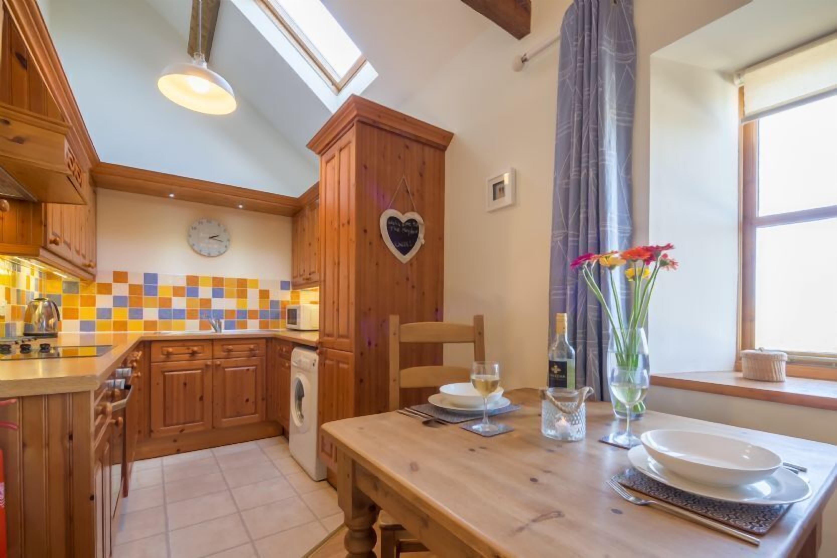 Una cocina con armarios de madera, salpicadero de azulejos y una mesa de comedor con platos, vasos y un jarrón de flores.