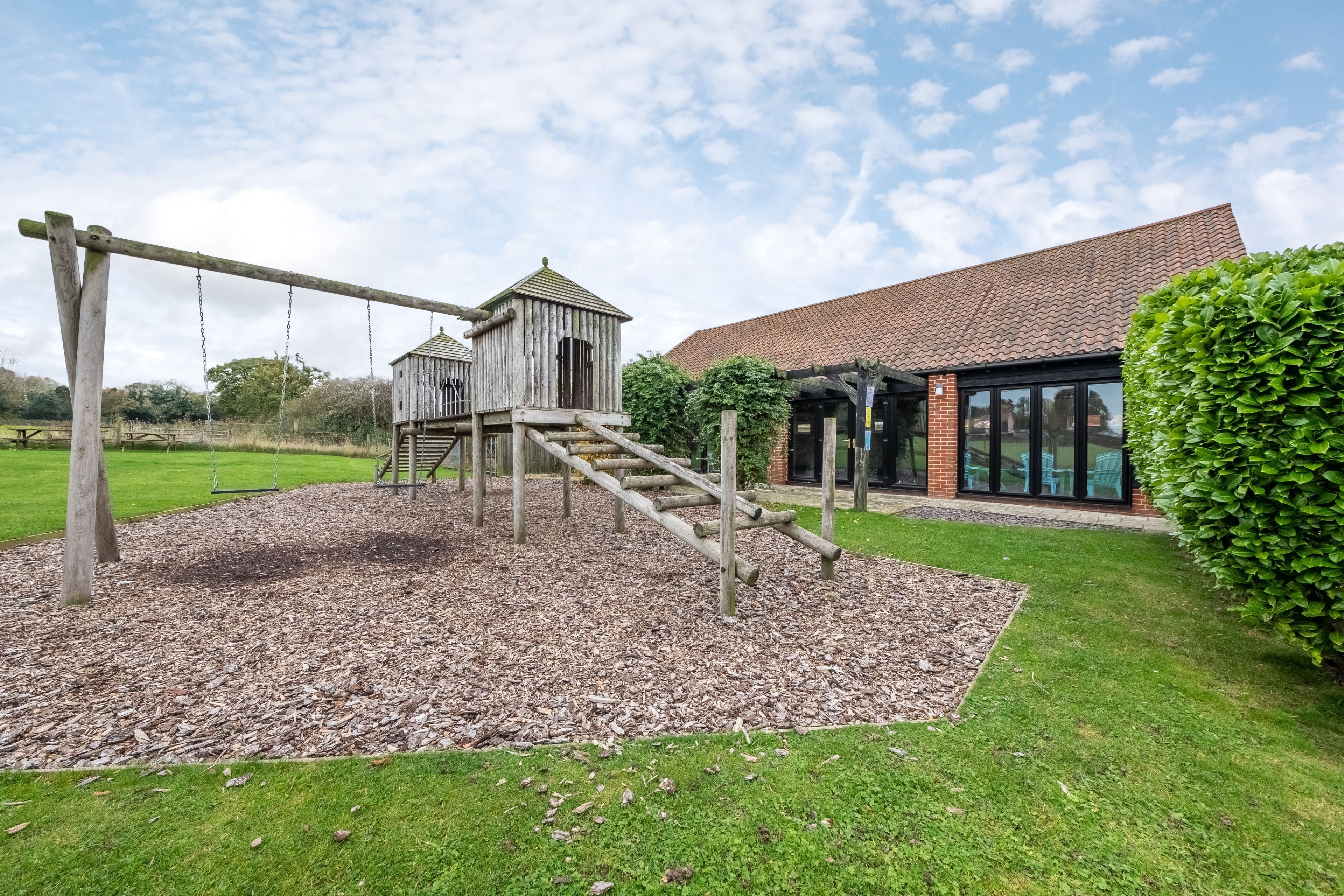 A playground with a wooden climbing structure and a small wooden building, surrounded by a grassy area and a building with a tiled roof.