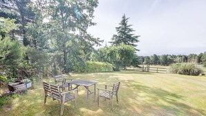 An outdoor seating area with a wooden table and benches, surrounded by trees and a grassy lawn.