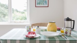 A breakfast table with a checkered tablecloth, a coffee pot, a kettle, a mug, a plate, a glass of orange juice, and a red and white cloth napkin.