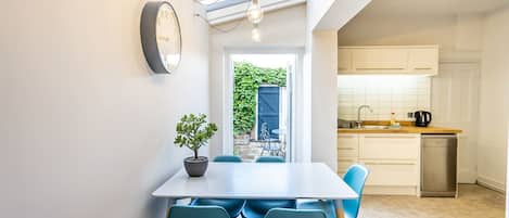 A modern kitchen with white cabinets, a sink, and a dishwasher. There is a dining area with a table and chairs, and a potted plant on the table.