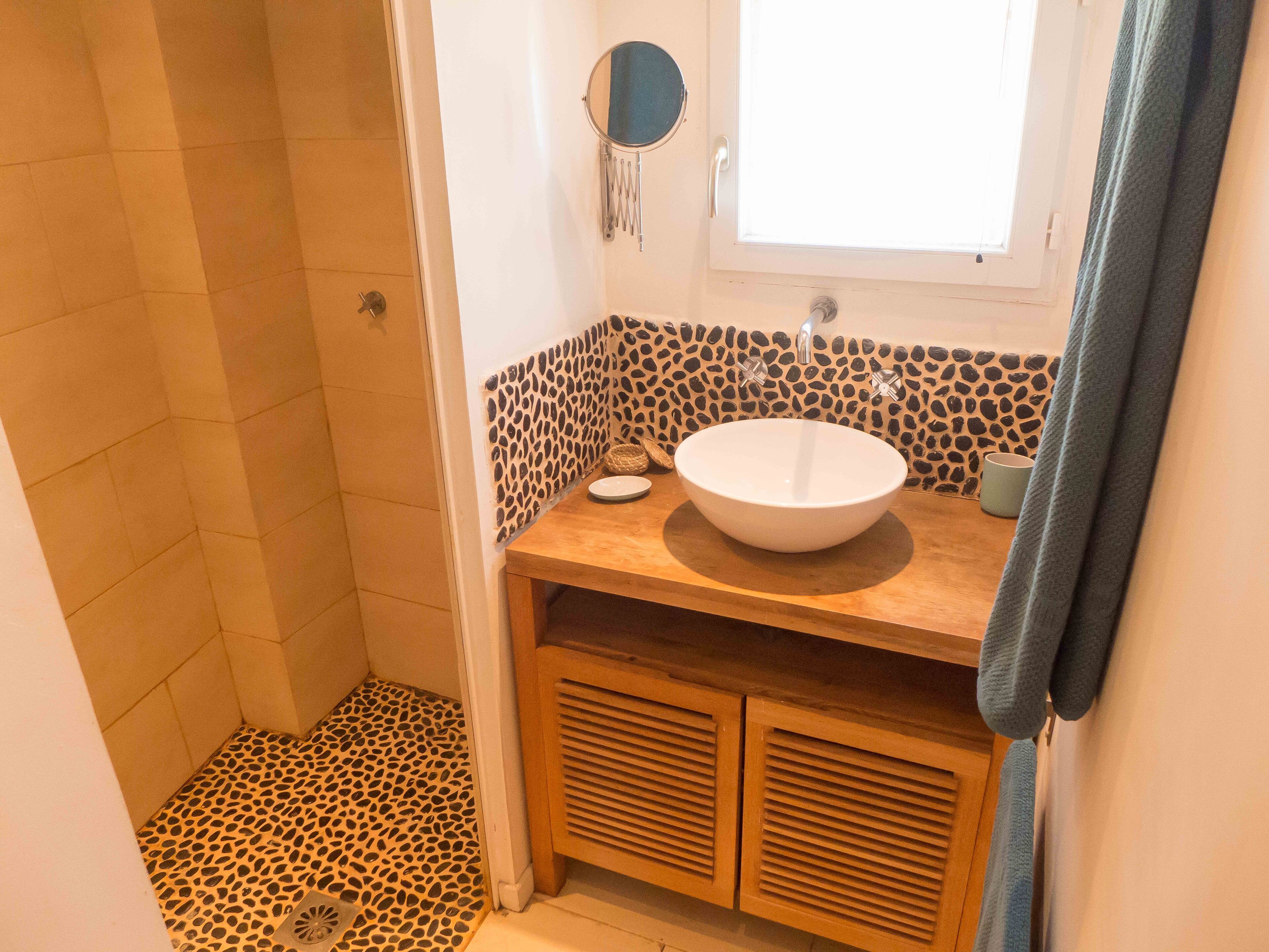 Un bagno con pavimento doccia effetto pietra, un mobiletto vanità in legno con lavandino bianco e uno specchio a cornice rotonda.