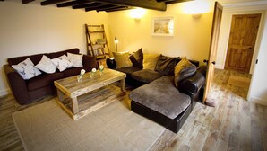 Un salon chaleureux avec un canapé marron, une table basse en bois et un canapé gris. Une échelle en bois est adossée au mur, et une porte donne accès à une autre pièce.