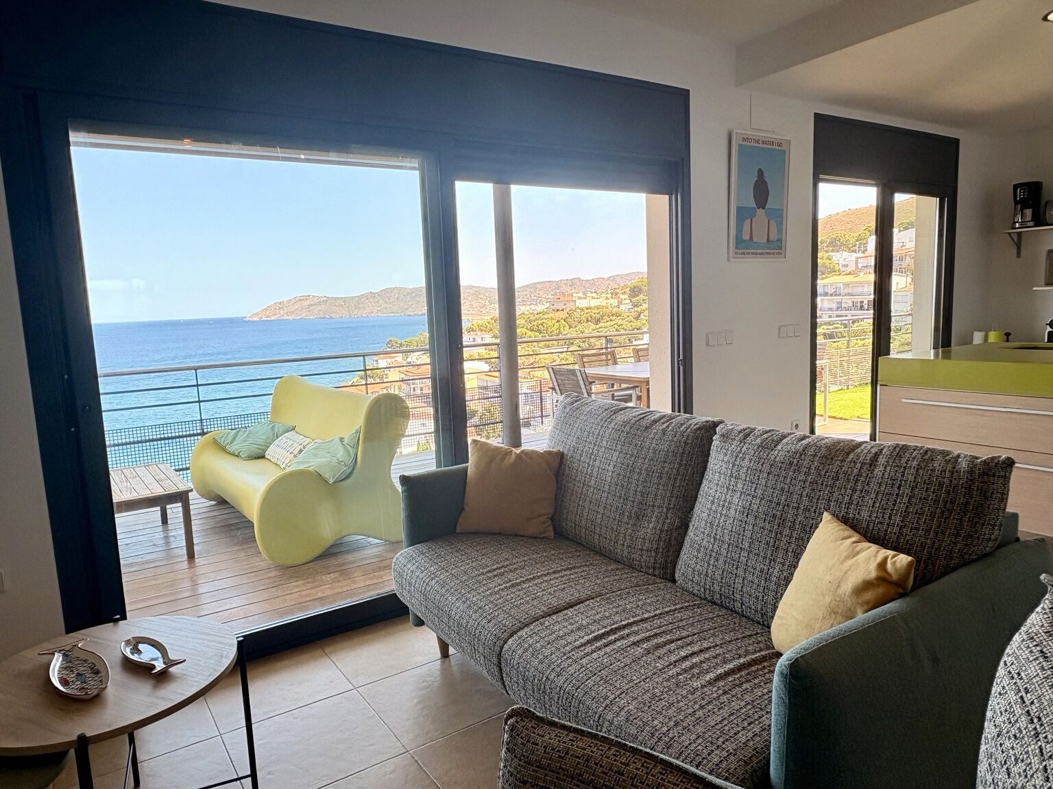 Una sala de estar con un sofá, un sillón amarillo y vista al mar a través de ventanas amplias.
