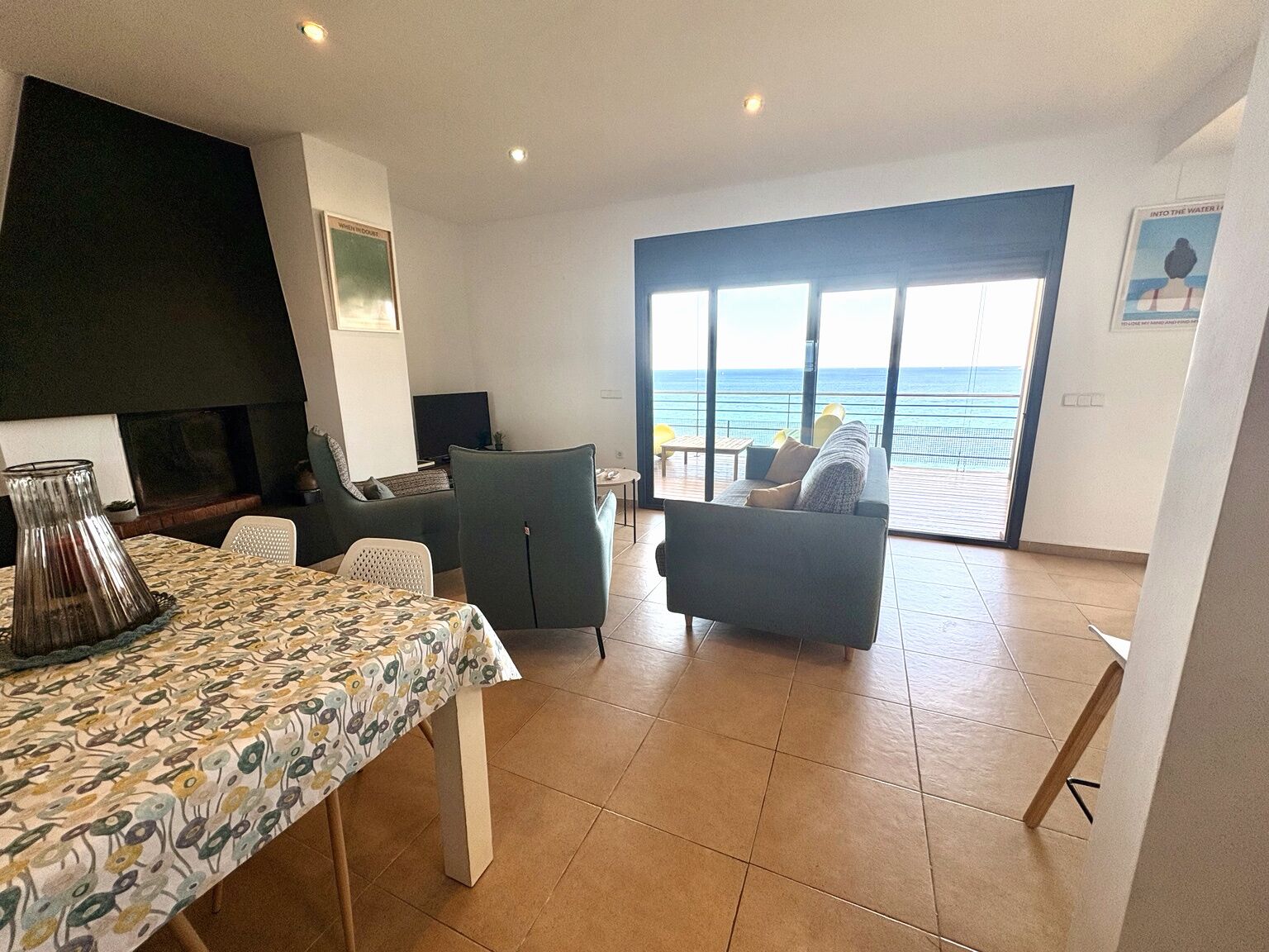 Uma sala de estar ampla com uma grande janela com vista para o mar, lareira e área de jantar com toalha de mesa estampada.